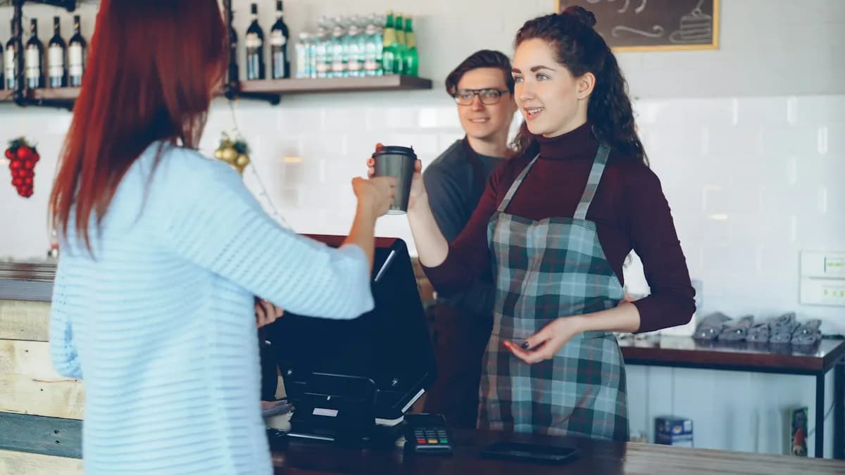 Kafe bankosunda iki görevli; biri müşteriye kahvesini uzatıyor, bankonun üzerinde POS terminali ve kart okuyucu duruyor